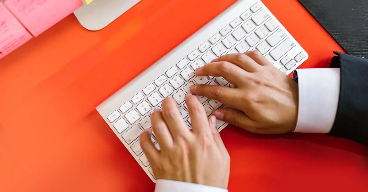 Gros plan sur des mains tapant sur un clavier sans fil sur un bureau orange, idéal pour des thèmes technologiques et professionnels.