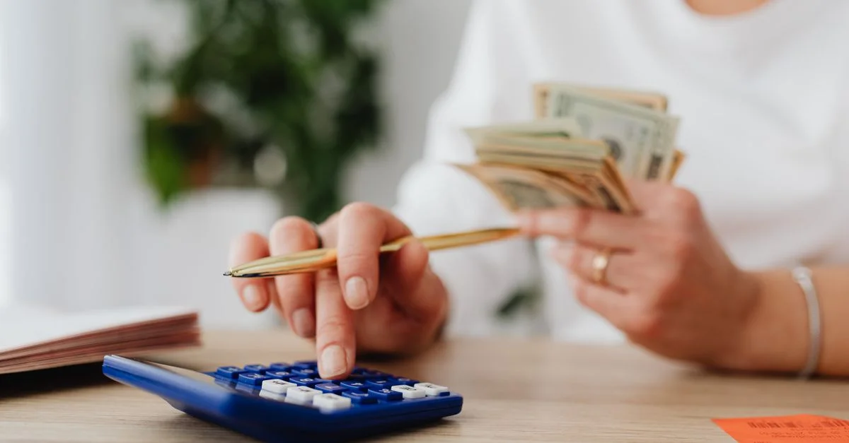 Une personne est assise à un bureau, calculant des finances à l'aide d'une calculatrice et tenant de l'argent.