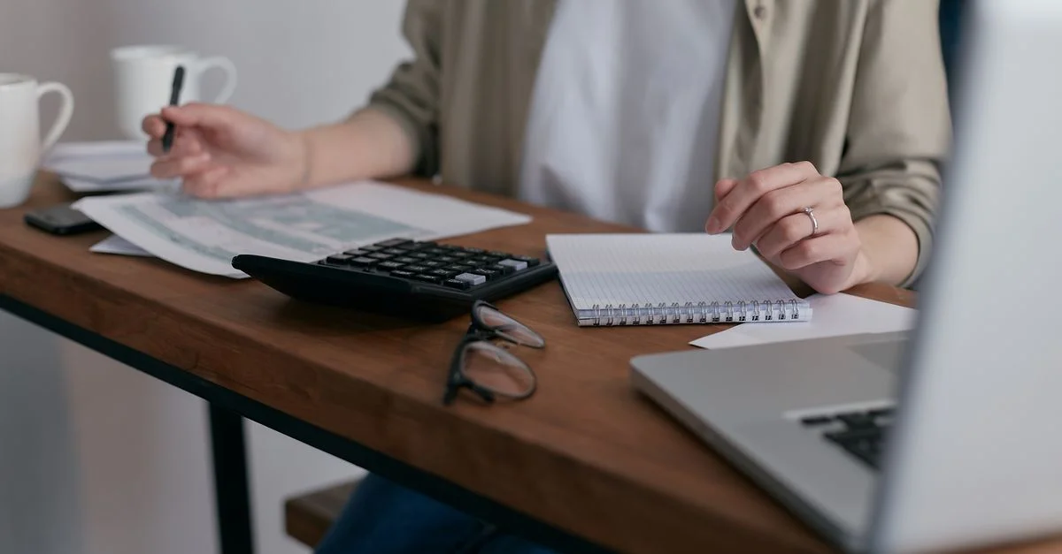 Une femme gère ses finances à domicile, utilisant un ordinateur portable et une calculatrice sur un bureau en bois.